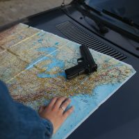 A handgun rests on a map on a car hood, symbolizing planning or escape.